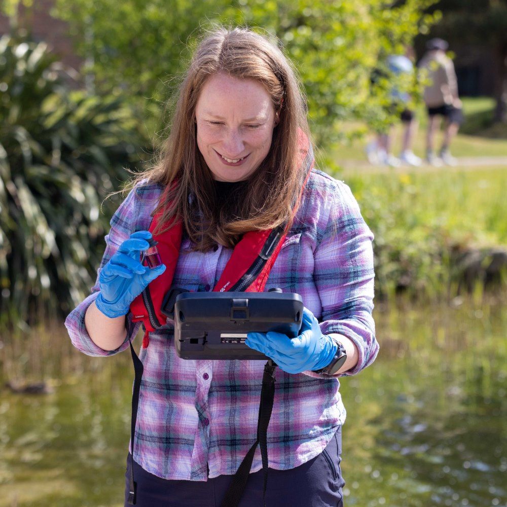 Dr Janet standing in a pond, wearing a purple checked shirt looking at a monitoring screen.