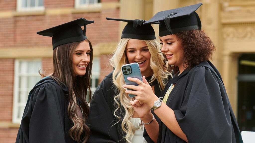 Three graduates wearing caps and gowns stood in front of the main building looking at a phone.