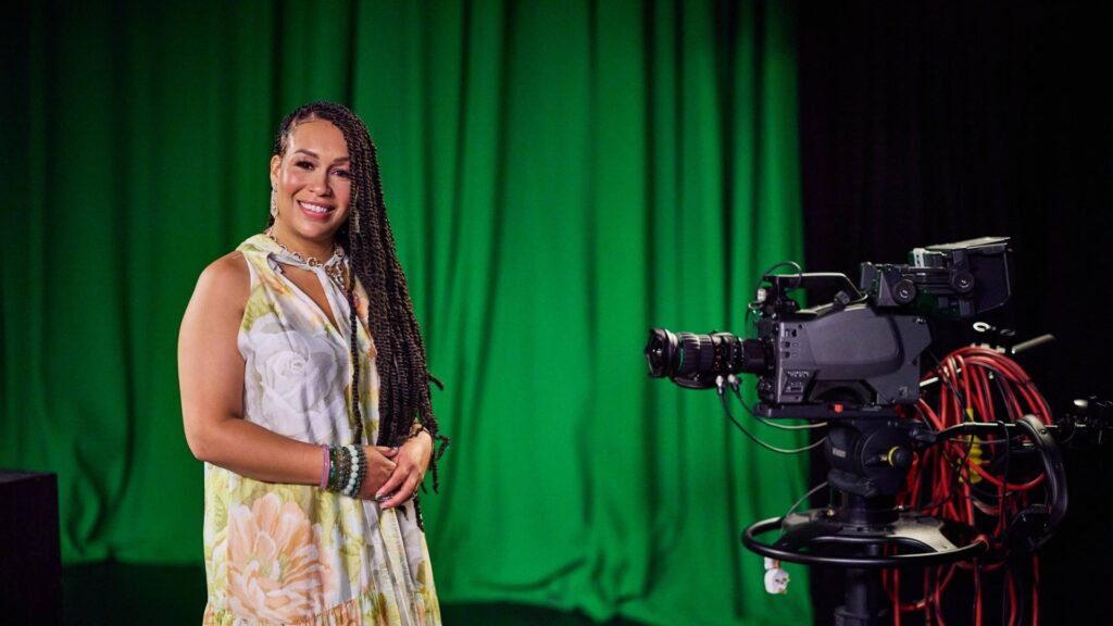 Rebecca Ferguson MBE inside tv studio with green curtain at the back and studio camera facing her, while she smiles at camera.