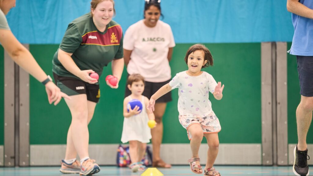 two little girls playing a game in the gym hall with two adults watching them.