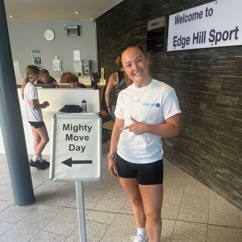 Faye Dunn, wearing black shorts and white tshort pointing to a sign that says Mighty Move with an arrow pointing to the left.