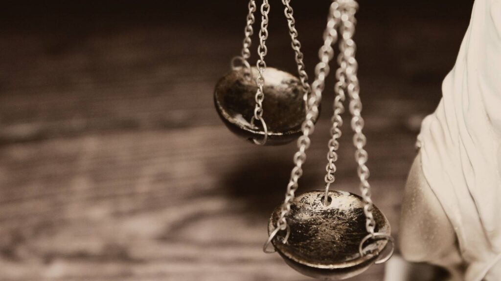 Close-up photo of scales of justice. One tray is in the foreground, the other the background, both suspended by chains. A part of a statue holding them is shown in the corner.