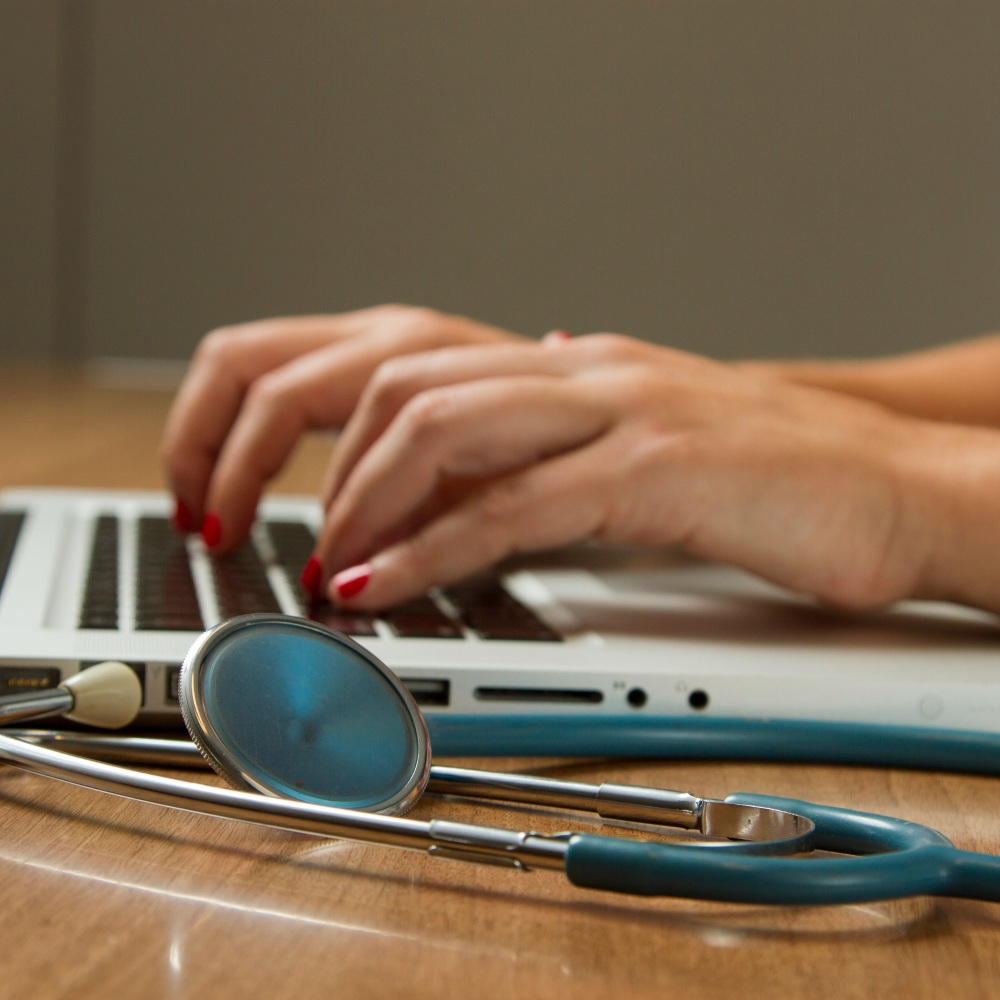 A pair of hands type at a laptop with a stethoscope in front.