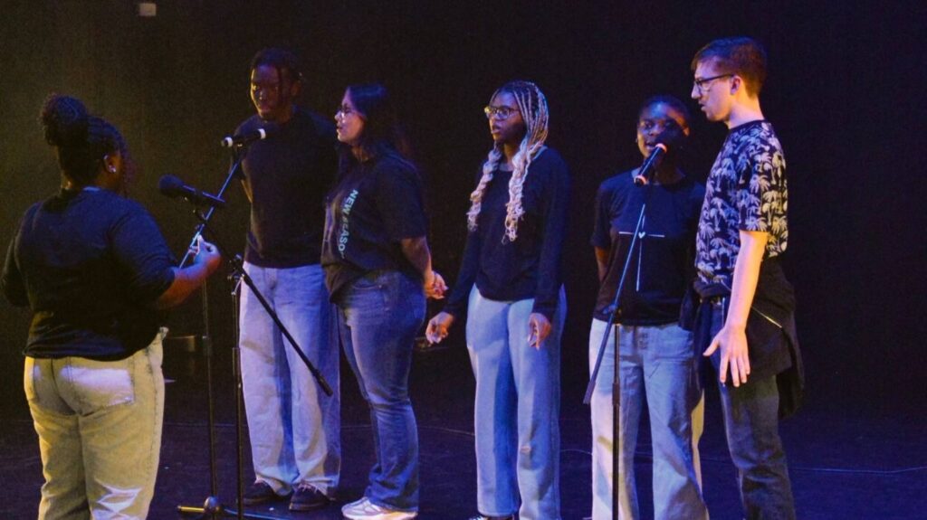 six students standing on a stage singing into a mic.