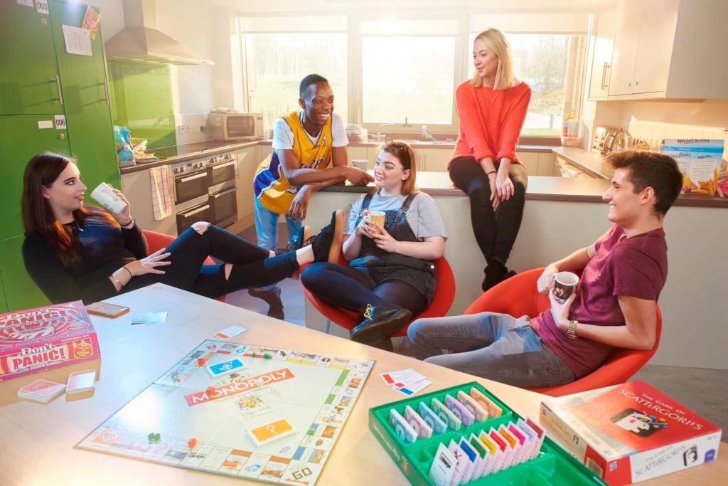 Five students chat in the kitchen and communal area of a halls of residence while drinking from mugs, with Monopoly and other board games on the table alongside them.