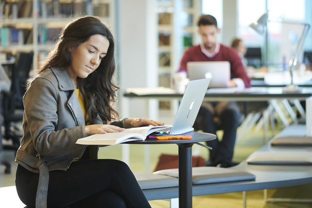 How to apply - A student reads a book while working on her Apple Macbook at a table in the University library in the Catalyst building.