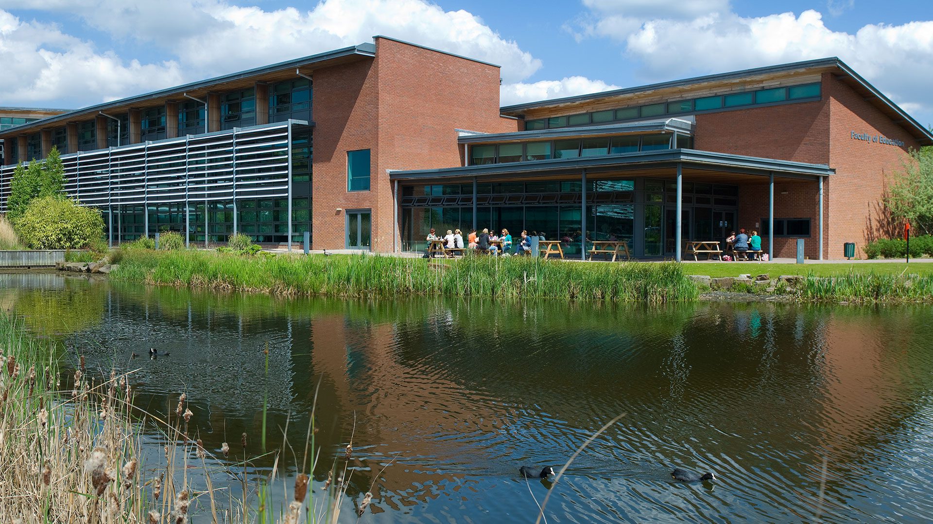 The lake in the foreground with the Faculty of Education building in the background. Ducks swim on the lake, and people sit outside the Faculty of Education on picnic benches.