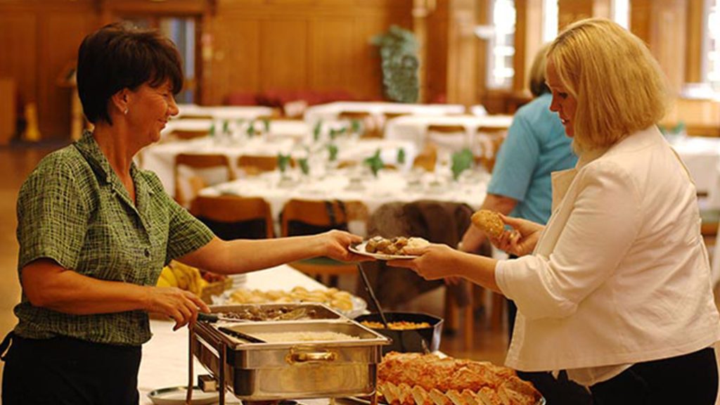 A member of staff hands someone a plate of food, in the background the room is set up for a dinner.