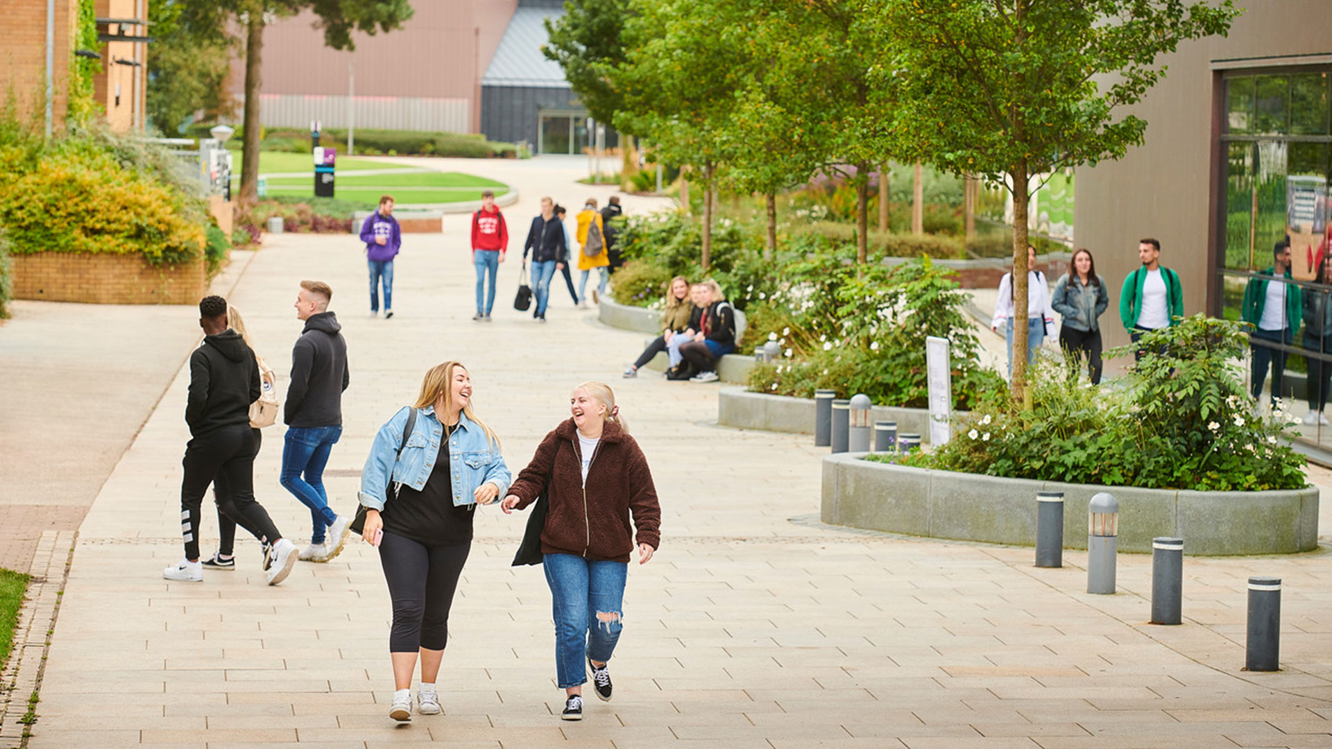 Two students walking together whilst laughing through campus. There are other groups of students walking together in the background.