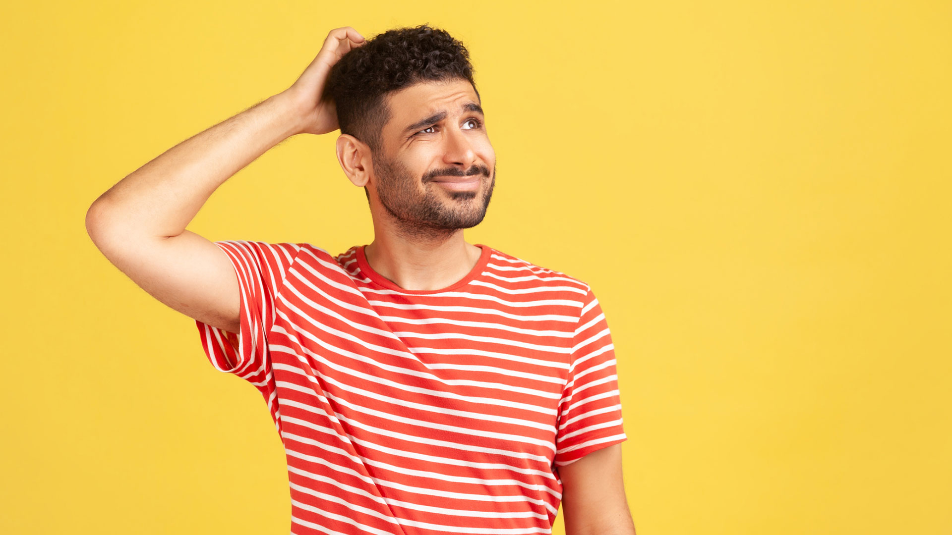 Confused uncertain man with beard in red striped t-shirt scratching his head, choosing, trying to make right decision, dilemma. Indoor studio shot isolated on yellow background