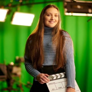 Professional Image of Amelia Cornett. Amelia has long red hair and is smiling for the camera whilst holding a film clapper.