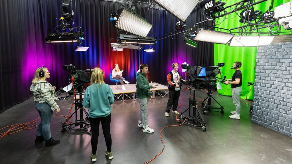 Students operate cameras in the TV studio in Creative Edge, filming presenters on a stage behind them.