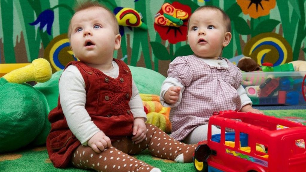Two babies sitting in a playroom surrounded by baby toys