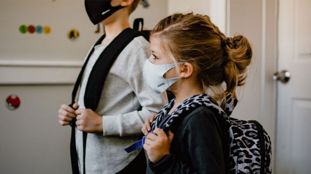 Two young children stood next to each other wearing face masks. They both are wearing a backpack and holding the straps.