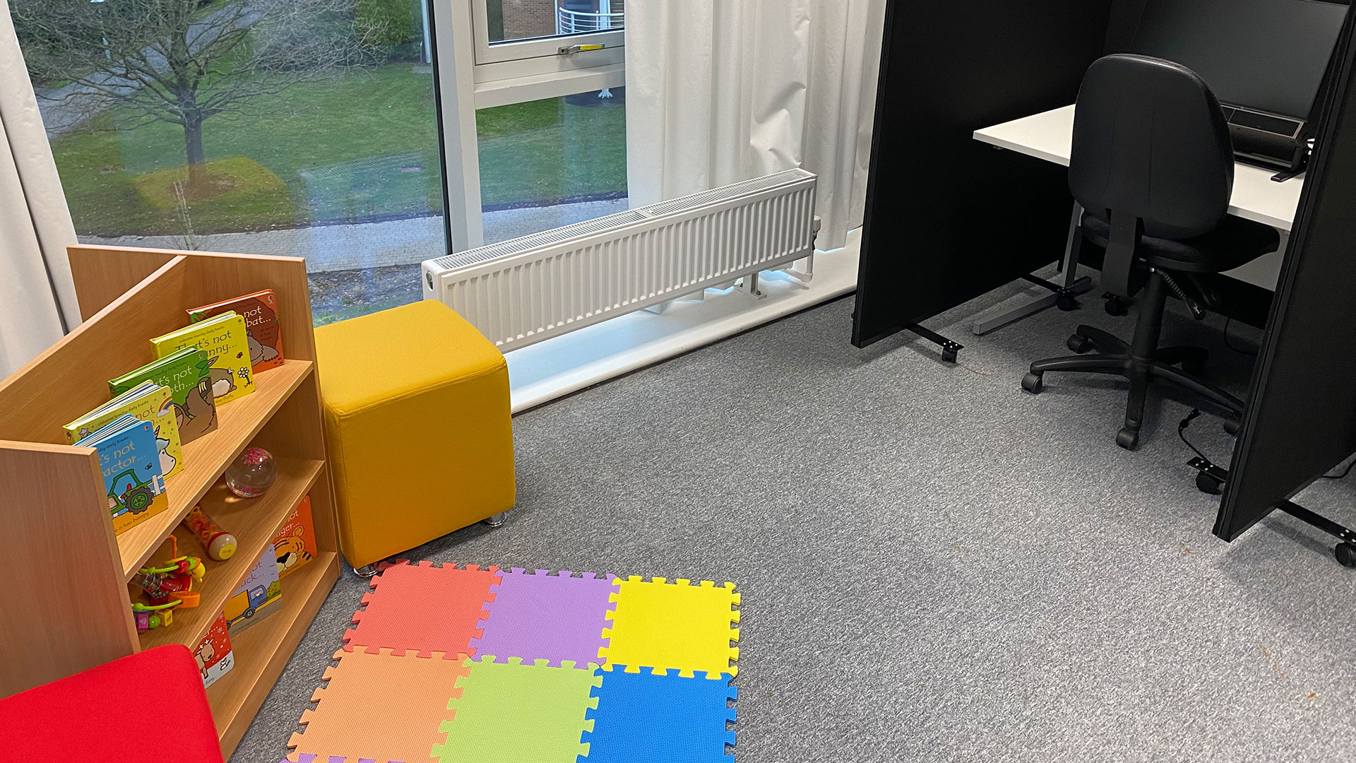 On-campus BabyLab room. There are two colourful stools at either side of a children's bookshelf. There is a child's soft play mat on the floor. To the right of the room there is a single person desk with a computer on it.