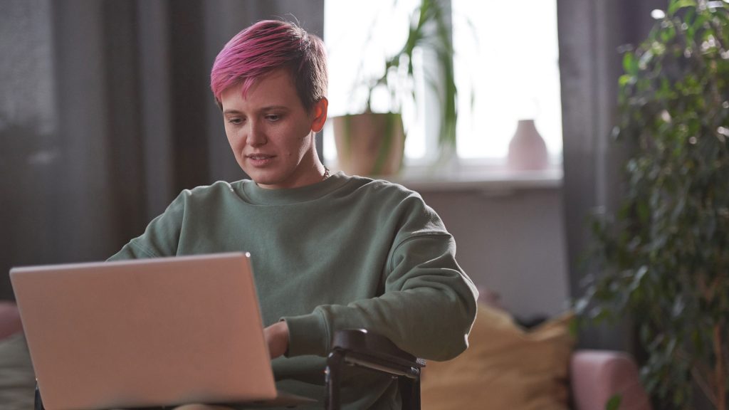 Young disabled woman sitting in wheelchair and typing on laptop computer. She has short hair that is dyed hot pink.