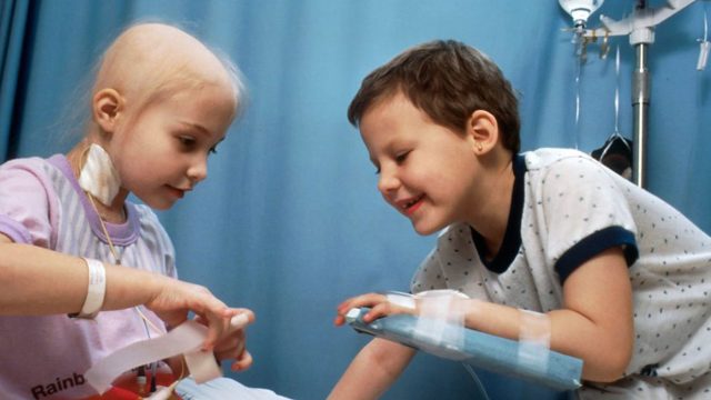 Children in hospital, one with a splint on their arm and the other with a tube coming out of their neck. They are smiling together on a hospital bed.