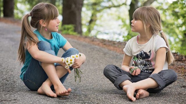 Two young children sitting on the floor outside, talking to eachother. One is holding a handful of daisies.