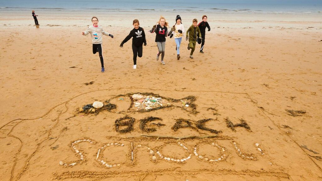 A group of children running towards the words "BEACH School" written in the sand using shells, stones and other items found on a beach.