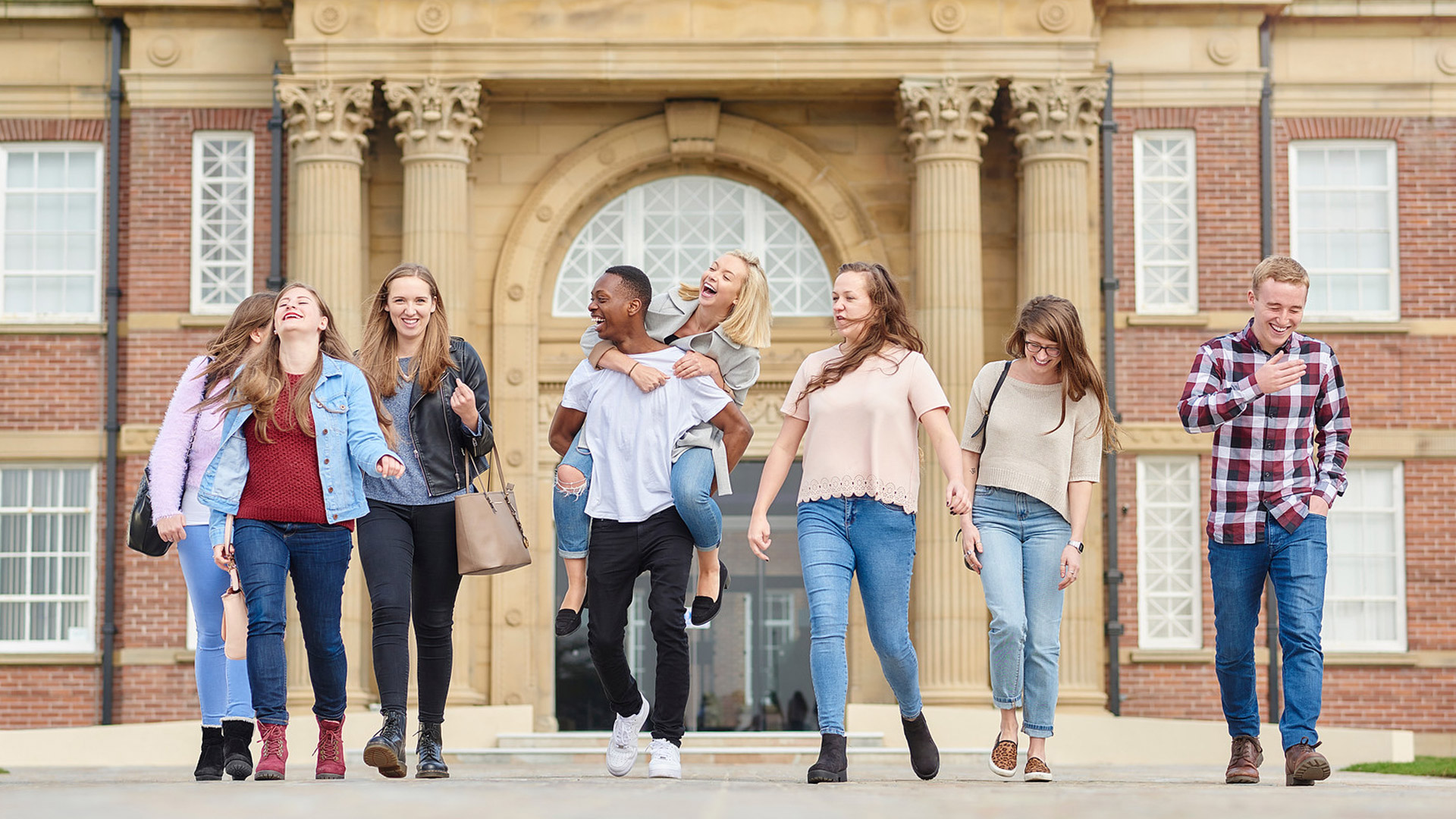 A large groups of students walk and laugh together outside of the main building.