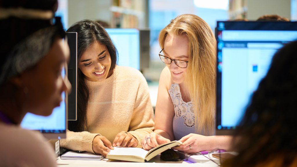Students looking at a text book while sat in front of a computer