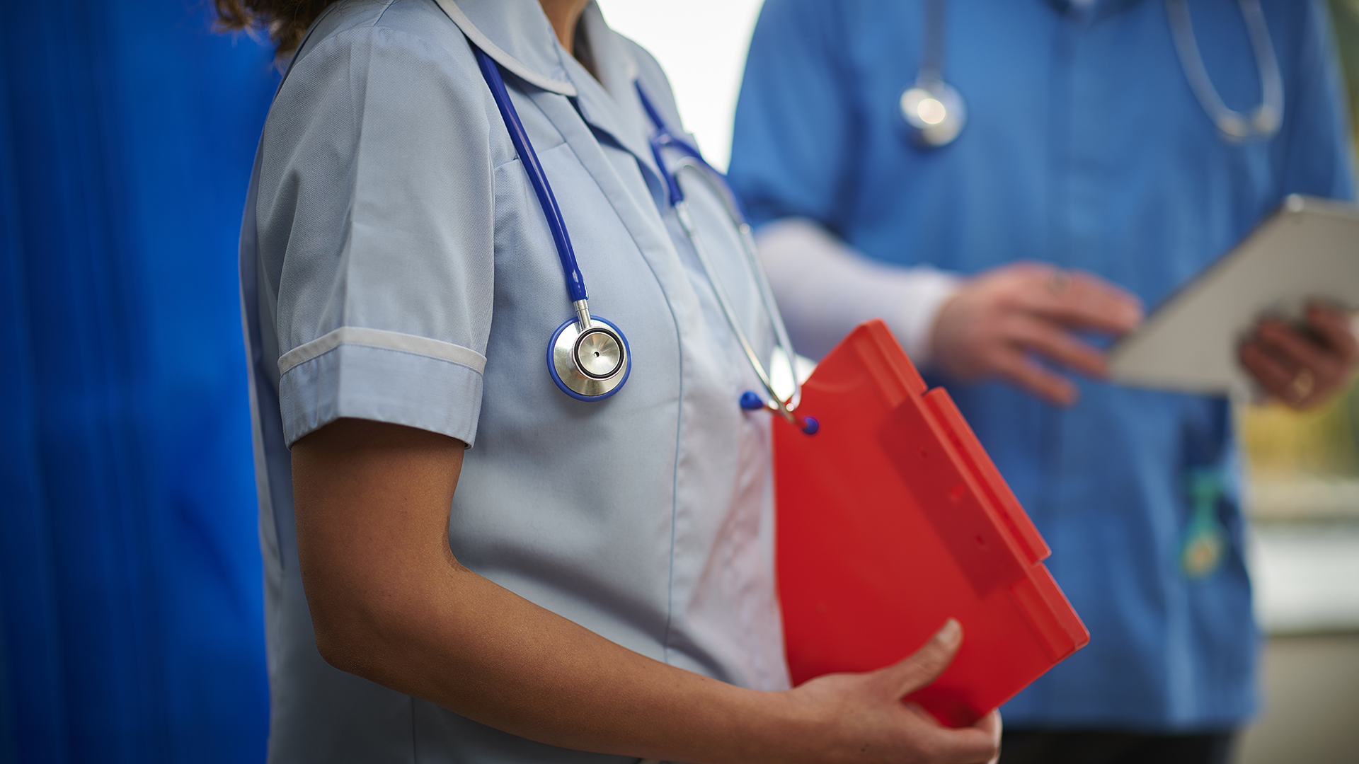 Mid body shot of a nurse holding a red clip board dressed in Edge Hill's pale blue nursing uniform.