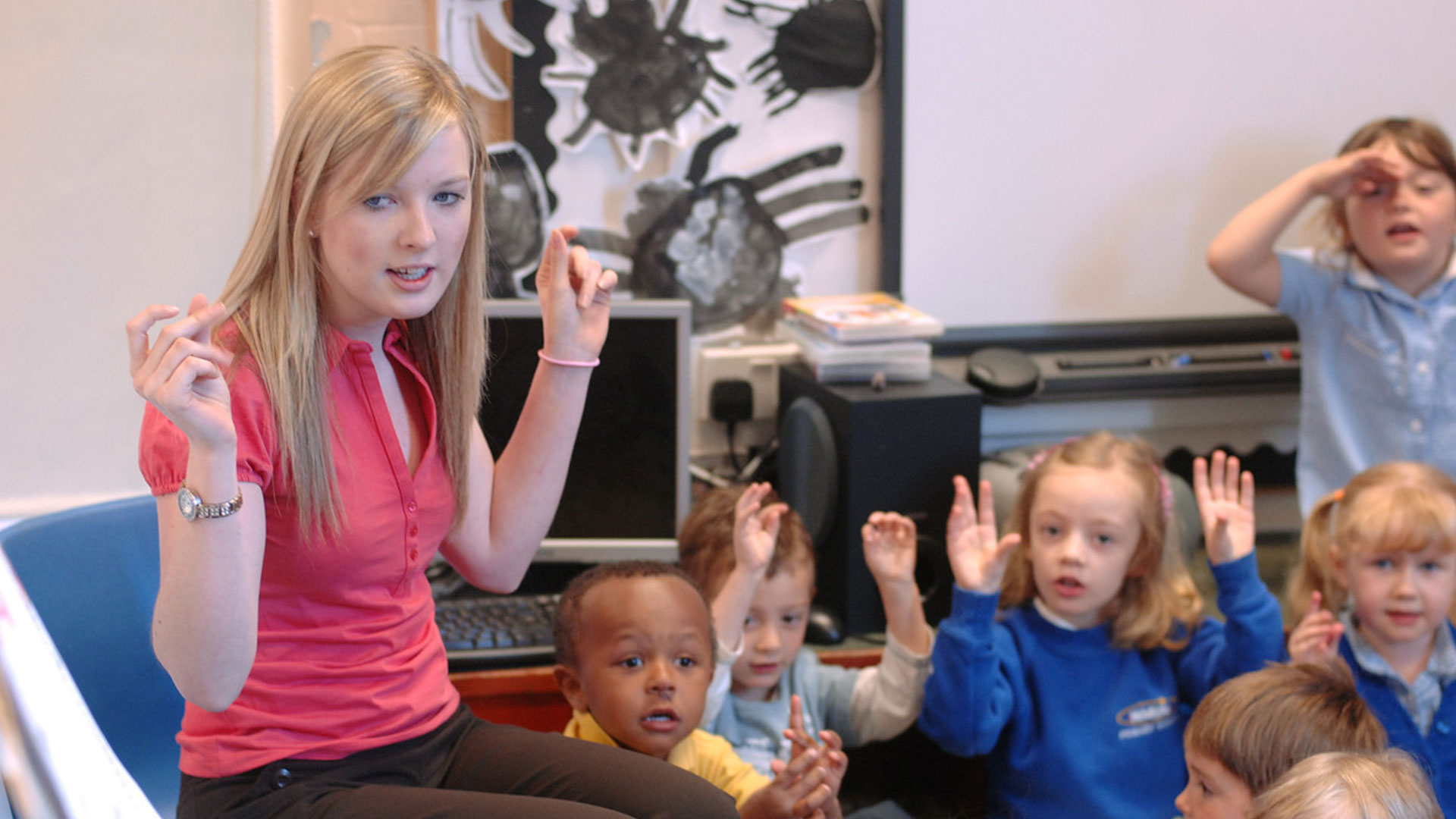 A teacher sits on a chair, singing a song with young children sitting on the floor