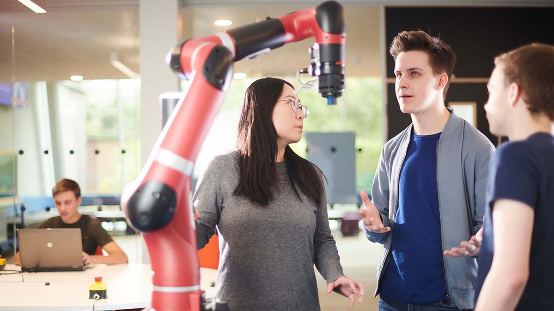 Three students chat in front of a robotic arm