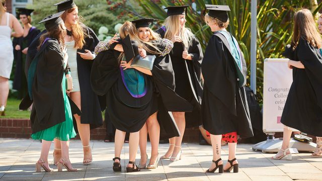 A group of graduates in their cap and gowns on their graduation day. The graduates are stood together in a group and two of them are hugging.