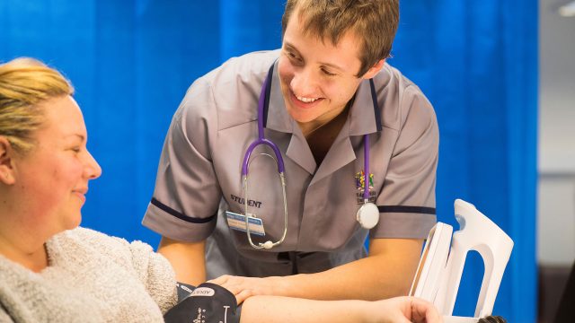 Health student with a stethoscope round their neck taking the blood pressure of a patient in a clinical setting