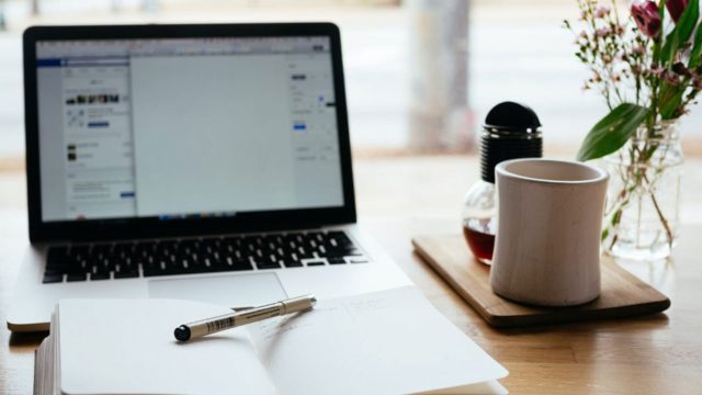 A laptop with an open notepad and pen, next to it is a mug and a vase of flowers