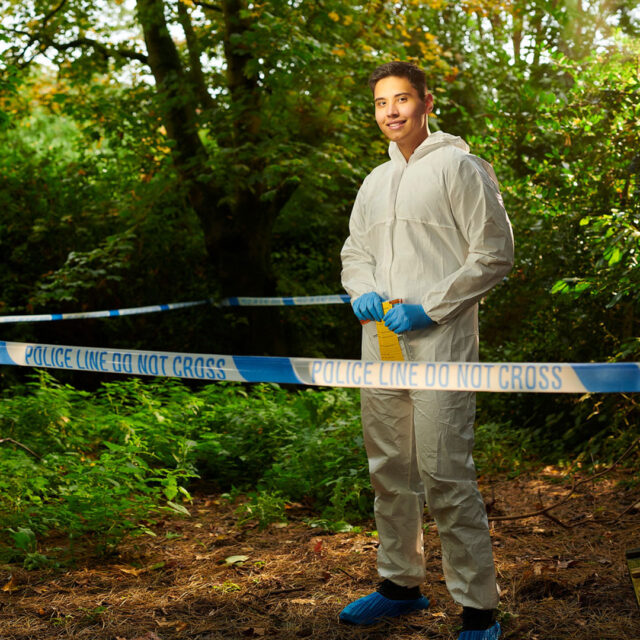Policing student, Luke Tomlinson. Luke is standing simulated crime scene setting. The setting is an outdoor woodland area taped up with police taping. Luke is wearing a forensics suit and rubber gloves.