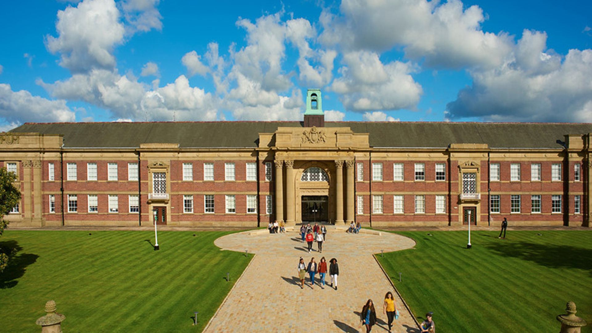 Exterior shot of the front of the main building on a sunny day. There are students walking down the path towards the building.