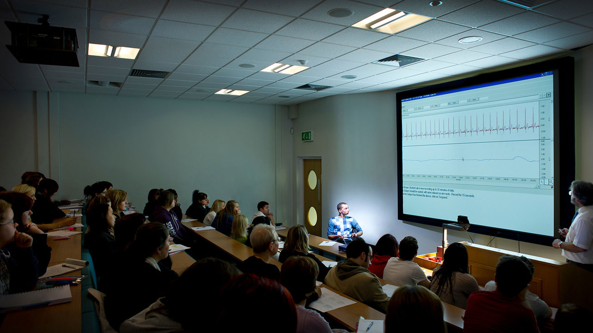 Two academics teaching a lecture theatre full of students. There is a smartboard displaying lecture content that they are looking at.