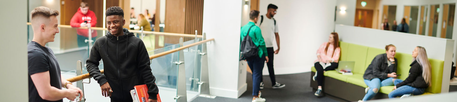 Students in the Law and Psychology building. There are two students leaning on the indoor balcony railing whilst smiling at each other. There are other students in the background. Three students sit on a lime green couch in the background and two other students are stood in front of the couch talking with them.