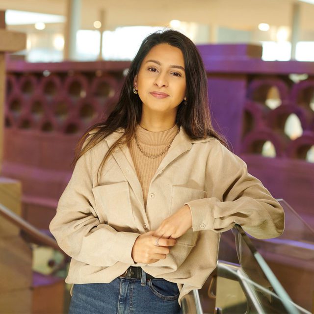 BA (Hons) English Literature and History student Sadiya Mansur leaning on the glass bannister in the Hub. There is a purple ambient glow in the background.