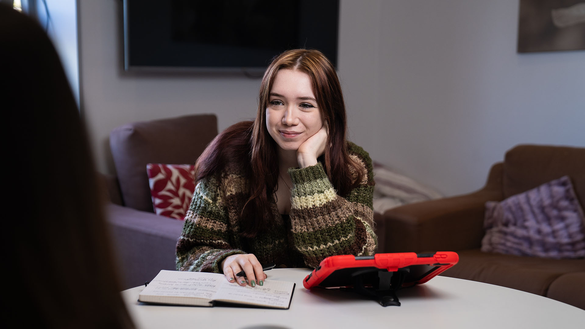 A student sits at a table with a notebook and tablet, listening and taking notes