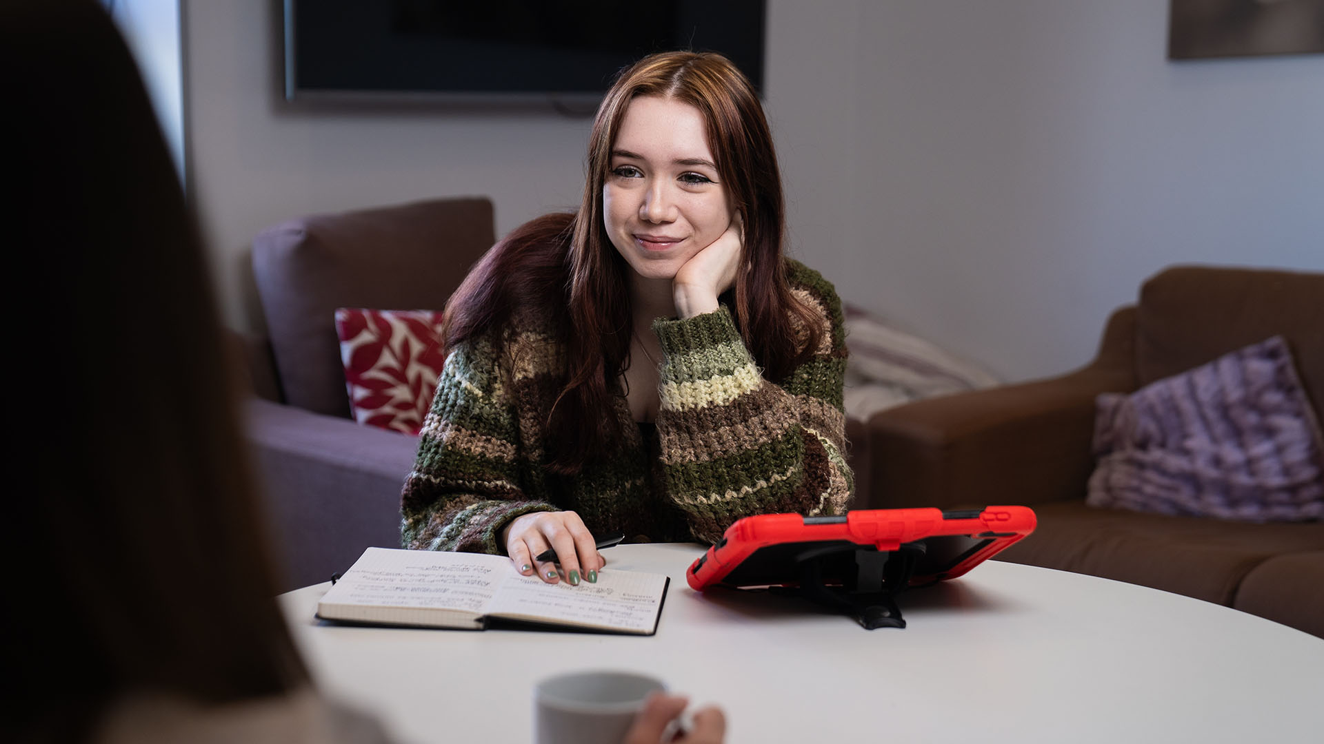 A student sits at a table with a notebook and tablet, listening and taking notes