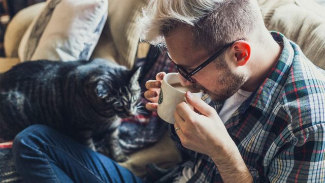 A man drinking a cup of tea with a cat next to him
