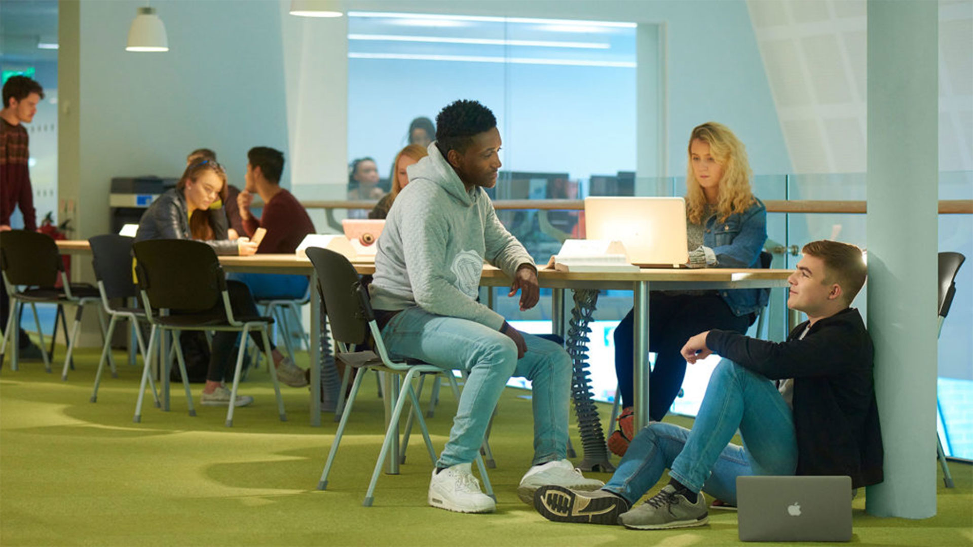 A group of students sit together. Two at a table, one using a laptop. The other person is having a conversation with another student who is sitting on the floor.
