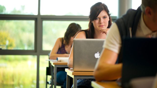 Students sitting in a classroom, focused on one student who is looking at a laptop