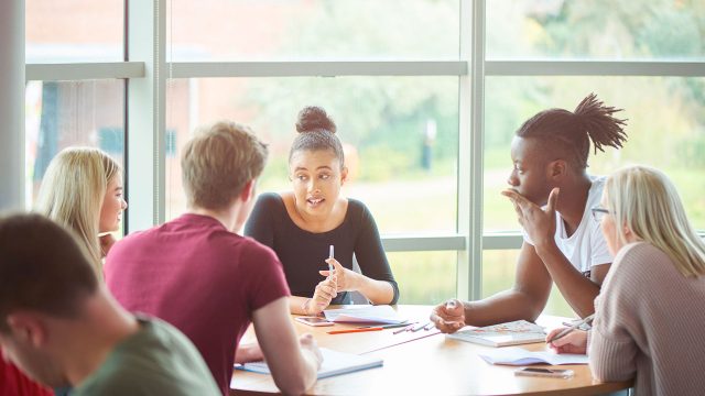 Students sitting around a table in a classroom, talking to each other with pens and paper on the table