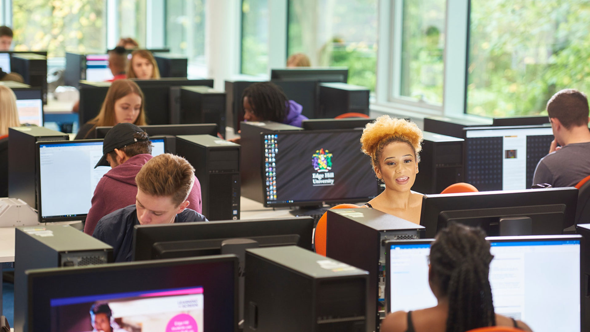Students in a computer classroom. Each student is at their own computer and one student is talking to another across the desk.