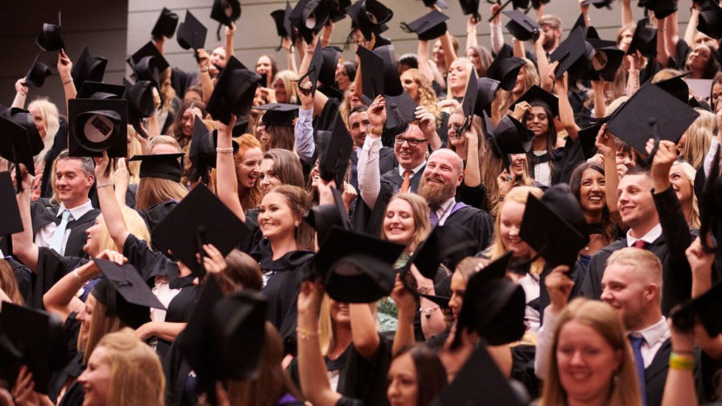 Students at graduation wearing black gowns and waving their mortar boards in the air