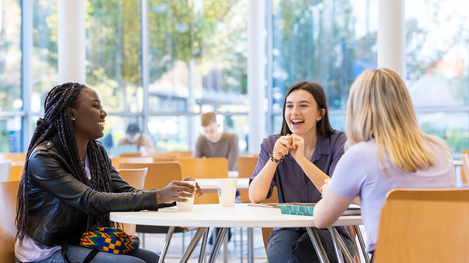 Three students sat at a table in the Hub, whilst laughing together. Two of the students have Starbucks drinks.