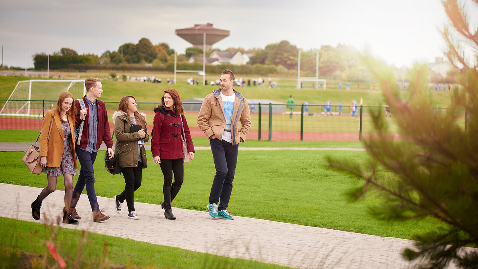 Five students walking on a path on campus. There are students in the background playing football on the sports pitch.