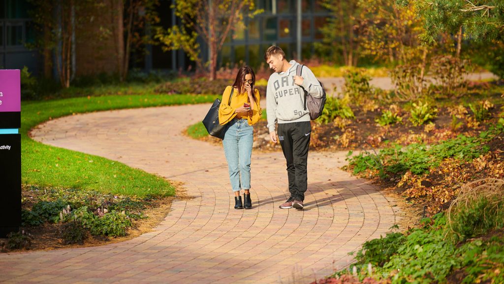 Two students walking down a path together on campus. One student of the students is on their phone, showing the other student what is on their screen.