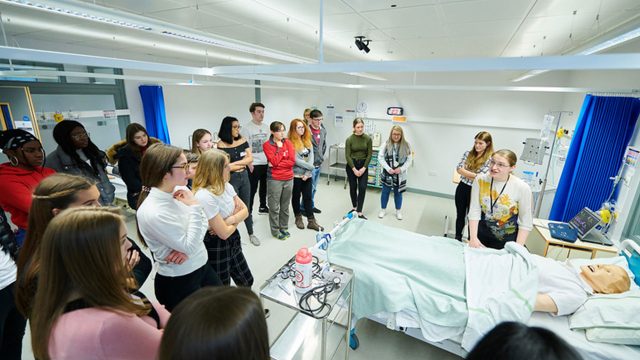 Students crowded round a hospital bed in the clinical skills and simulation centre, watching someone who is talking to them