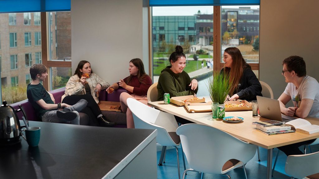 Three students sat at their dining table in their student accommodation, eating takeaway pizza. Three other students are sat behind them on a couch, eating takeaway pizza. There are large windows in the background showcasing the campus grounds, including the Catalyst building.