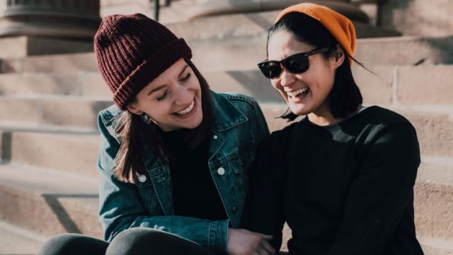 Two people sitting outside on some steps laughing together. They are both wearing beanies.